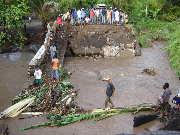 Dozens of people on the road looking at a bridge washed away by floods. 